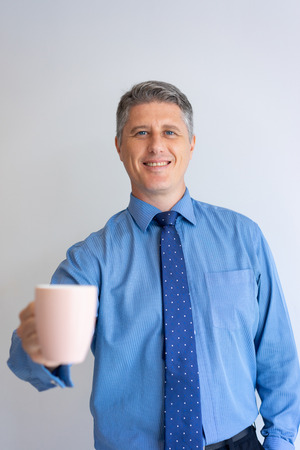 Happy business professional enjoying office morning. Mid adult Caucasian man in shirt and tie showing white cup at camera. Coffee break conceptの写真素材