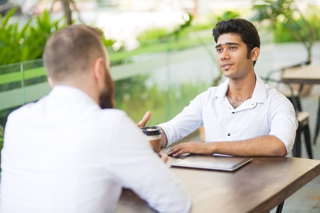 Confident handsome Indian manager talking to business partner at informal meeting in outdoor cafe. Serious young businessman asking colleague about project. Discussing business conceptの写真素材