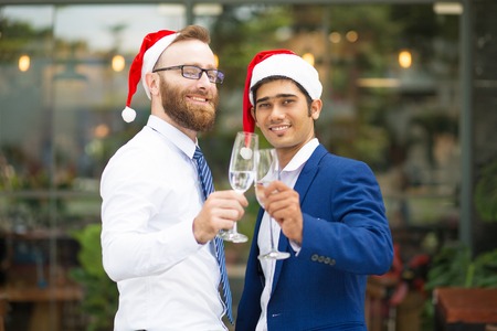 Happy excited multi-ethnic men clinking champagne flutes and looking at camera. Cheerful optimistic managers in formalwear celebrating Christmas. Office party conceptの写真素材