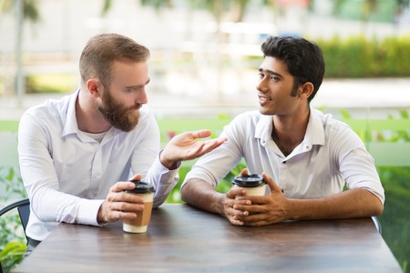 Two male friends drinking coffee and chatting in outdoor cafe. People sitting at table with blurred plants in background. Coffee break concept. Front view.の写真素材