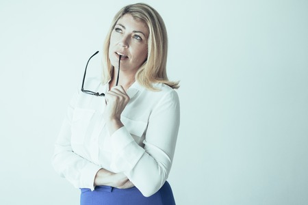 Closeup portrait of pensive middle-aged attractive fair-haired woman thinking and biting glasses temple. Contemplation concept. Isolated view on grey background.の写真素材