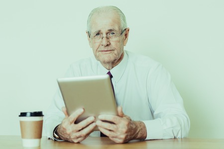 Closeup portrait of serious senior business man looking at camera and using tablet computer at office desk. Senior entrepreneur concept. Isolated front view on white background.の写真素材
