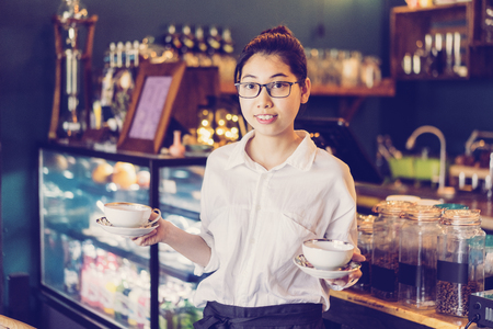 Cheerful pretty Asian waitress carrying coffee cups and looking at camera. Smiling attractive young coffee shop owner working in her cafe. Small business conceptの写真素材