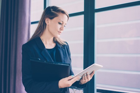 Closeup portrait of serious young beautiful brown-haired business woman using tablet computer and holding folder at window. Business communication concept.の写真素材