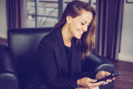 Closeup portrait of smiling young beautiful brown-haired woman using smartphone and sitting in armchair in hotel lobby. Business communication concept. Side view.の写真素材