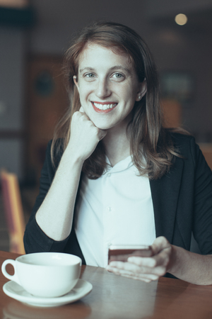 Portrait of young Caucasian woman, businesswoman or student, networking on smartphone and smiling at cafe. Coffee break conceptの写真素材