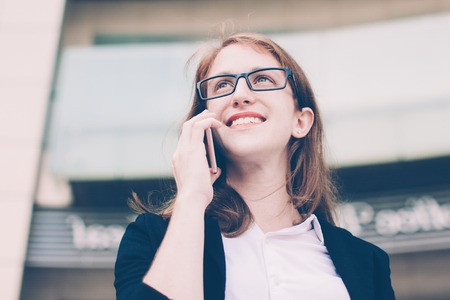 Portrait of happy young Caucasian businesswoman or student wearing glasses standing outdoors and talking on mobile, phone. Woman in business and mobile communication conceptの写真素材