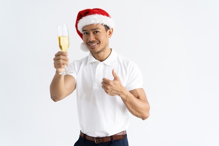 Asian man wearing Santa hat and raising goblet with champagne. Smiling guy showing thumb up. Christmas party and success concept. Isolated front view on white background.の写真素材