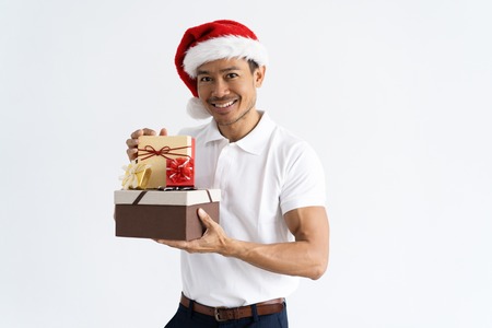 Happy man wearing Santa hat and showing gift boxes. Asian guy looking at camera. Christmas gifts concept. Isolated front view on white background.の写真素材
