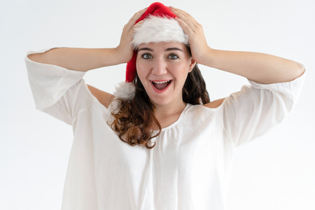 Excited Xmas girl overjoyed with amazing news. Beautiful young woman in Santa hat holding head and smiling with open mouth. Christmas surprise conceptの写真素材