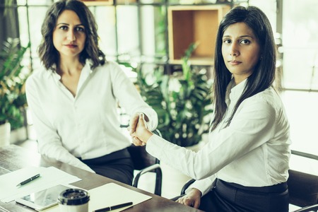 Two serious business women shaking hands and sitting at table in cafe. They are looking at camera. Business agreement concept.の写真素材
