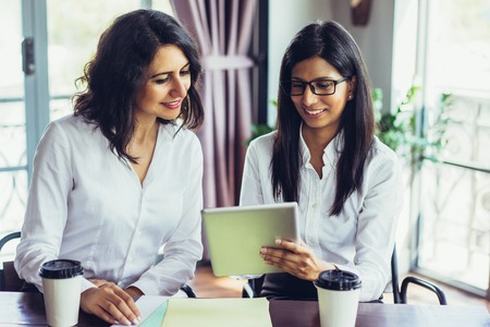 Smiling female business colleagues examining data on touchpad. Young Caucasian and Latin American businesswomen sitting at table and using digital tablet in office. Teamwork conceptの写真素材