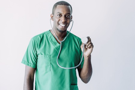 Portrait of cheerful young doctor listening with stethoscope. African American cardiologist wearing green medical garment looking at camera and smiling. Treatment and health conceptの写真素材
