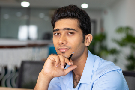 Portrait of young Indian businessman or student sitting with pen and looking at camera in office. Businessman conceptの写真素材