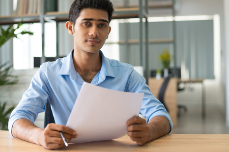 Content confident handsome Indian businessman analyzing data while examining report in office. Serious young business expert working with document. Paperwork conceptの写真素材