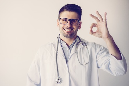 Portrait of cheerful male general practitioner showingok sign. Young Caucasian doctor wearing glasses, lab coat and stethoscope looking at camera and smiling. Success conceptの写真素材