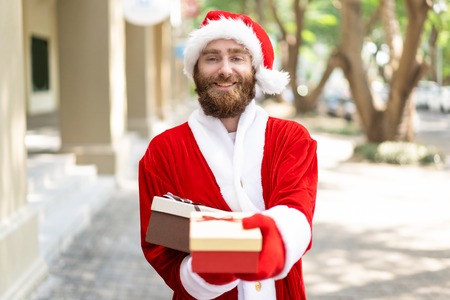 Cheerful Christmas hipster giving gifts. Bearded young man wearing Santa costume holding present in outstretched hand. Christmas gifts conceptの写真素材