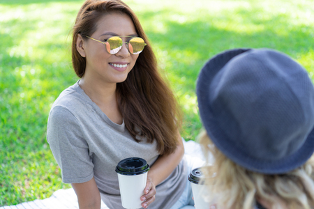 Smiling Asian girl with coffee cup chatting with close friend at picnic. Happy young woman in sunglasses enjoying leisure with friend outdoors. Communication conceptの写真素材