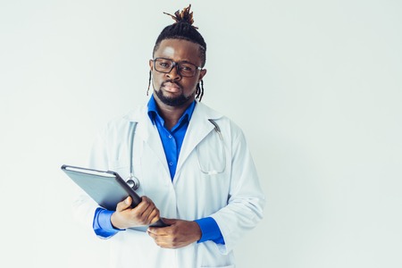 Serious calm young black male doctor in glasses holding folder and looking at camera. Confident medical specialist in lab coat standing against white wall. Hospital conceptの写真素材