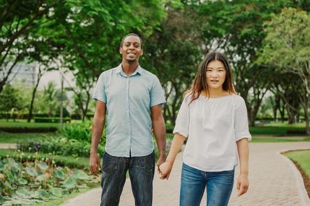 Happy Asian woman and Afro American man walking together holding hands in park. Asian woman and African American man dating in summer. Relationship and break conceptの写真素材