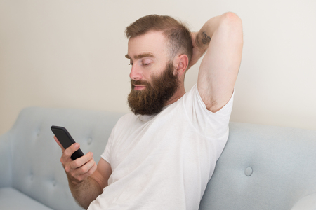 Focused man browsing on smartphone and sitting on sofa. Handsome guy keeping hand behind head and relaxing. Communication concept. Front view.の写真素材