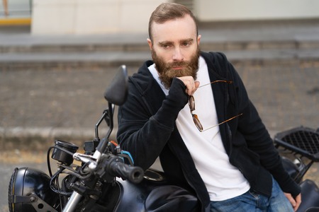 Portrait of pensive man standing at motorbike with sunglasses. Young Caucasian biker posing at his...