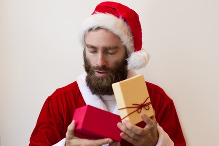 Serious guy wearing Santa costume and looking into gift box. Handsome bearded man. Christmas surprise concept. Isolated front view on white background.の写真素材