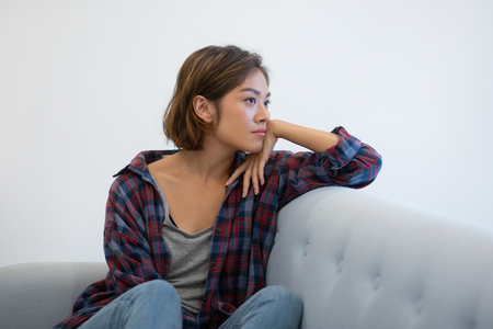 Pensive Asian girl on couch thinking over problems. Beautiful young woman in casual checkered shirt leaning head on hand and looking into vacancy. Unemployment conceptの写真素材