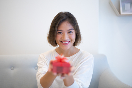 Cheerful young Asian woman giving Christmas present to you. Smiling attractive girl with short hair sitting on sofa in living room. Gift-giving conceptの写真素材