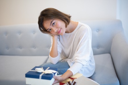 Content Asian woman in casual sweater holding Christmas gift and looking at camera. Calm girl sitting on comfortable sofa in living room. Celebration conceptの写真素材