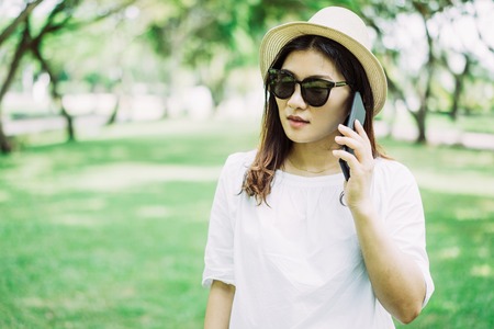 Serious pensive young Asian woman in sunglasses talking on mobile phone while walking in summer park. Pretty girl in straw hat strolling alone and using phone. Communication conceptの写真素材