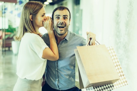 Young woman sharing good news with husband during shopping. Blonde woman in casual wear whispering in mans ear, shocked and happy man holding shopping bags. Shopping and surprise conceptの写真素材