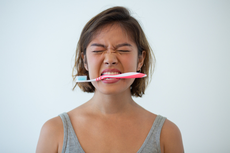 Portrait of mischievous young woman holding toothbrush in teeth. Frowning Asian girl during morning routine. Dental care conceptの写真素材