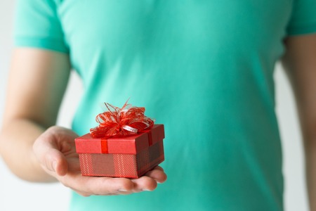 Closeup of guy holding small red gift box on palm. Man wearing t-shirt. Gift concept. Isolated cropped front view on white background.の写真素材