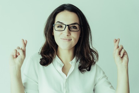 Positive cheerful female student in glasses making wish. Young Caucasian woman in white shirt showing fingers crossed. Luck and fortune conceptの写真素材