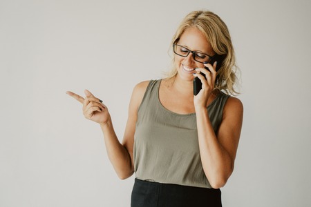 Happy blonde lady in glasses having nice phone call. Cheerful Caucasian woman speaking on mobile phone, laughing and pointing finger aside. Advertising and communication conceptの写真素材