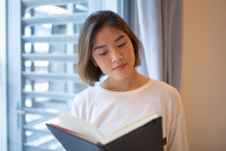 Focused young woman reading book at window at home. Pretty Asian lady holding book and standing at home. Education concept. Front view with window in background.の写真素材