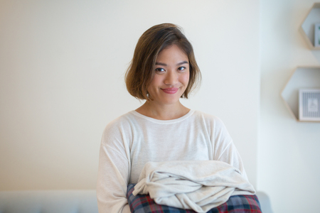Smiling pretty woman holding clean folded clothes at home. Young Asian lady looking at camera and standing with wall in background. Laundry and housekeeping concept. Front view.の写真素材