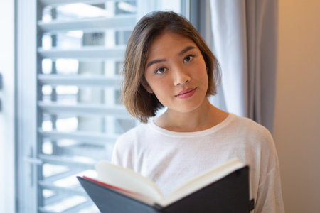 Serious young woman reading book at window at home. Pretty Asian lady looking at camera and standing at home. Education concept. Front view with window in background.の写真素材