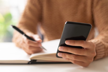 Close-up of lady writing out information from phone message. Unrecognizable businesswoman making notes in diary and viewing online file on smartphone. Gadget conceptの写真素材