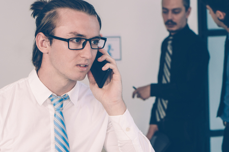 Serious young manager having phone call during corporate meeting. Long haired man in tie speaking on cellphone, his colleagues talking in background. Business communication conceptの写真素材