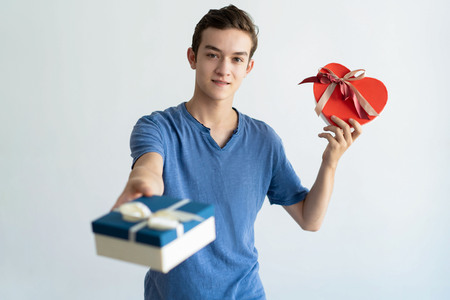 Smiling teen boy giving gift box to viewer. Young man standing, holding two gift boxes and looking at camera. Gifts and holiday concept. Isolated front view on white background.の写真素材
