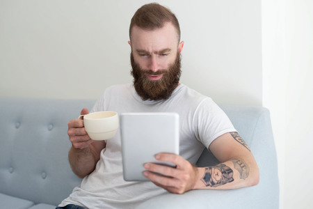 Excited bearded guy enjoying coffee break and video chat. Young man sitting on couch, holding cup...