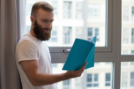 Serious hipster man in tshirt reading book. Concentrated young guy with beard standing at window....