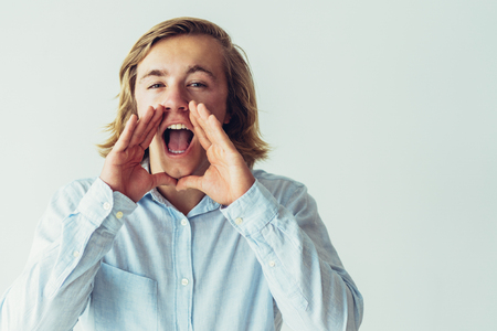 Cheerful handsome guy announcing important news. Young long haired blonde man keeping hands at mouth and shouting loud. Advertising conceptの写真素材