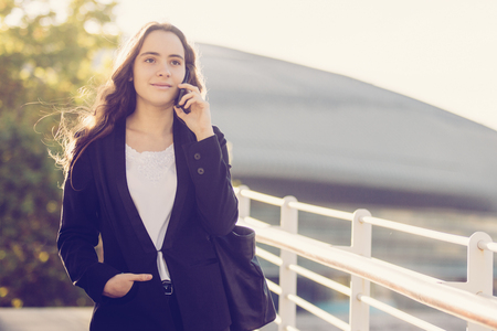 Smiling female assistance speaking on phone on her way to office. Beautiful young woman in casual jacket walking outdoors and talking on cellphone. Connection conceptの写真素材