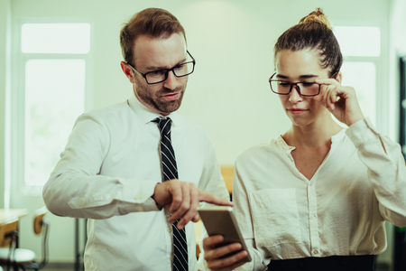 Serious business people reading news on smartphone. Woman and man standing in office with blurred interior in background. News concept. Front view.の写真素材