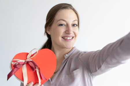 Cheerful lady taking selfie photo with heart shaped gift box. Woman posing and holding gadget which is out of view. Valentines Day and technology concept. Isolated front view on white background.の写真素材