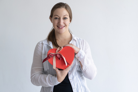 Happy pretty young woman holding heart shaped gift box. Girl looking at camera. Saint Valentines Day gift concept. Isolated front view on white background.の写真素材