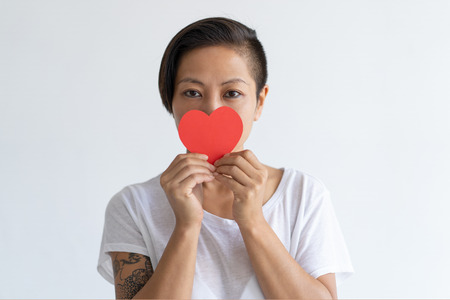 Playful Asian woman holding paper heart in front of her mouth. Tattooed lady wearing t-shirt and looking at camera. Saint Valentines Day concept. Isolated front view on white background.の写真素材